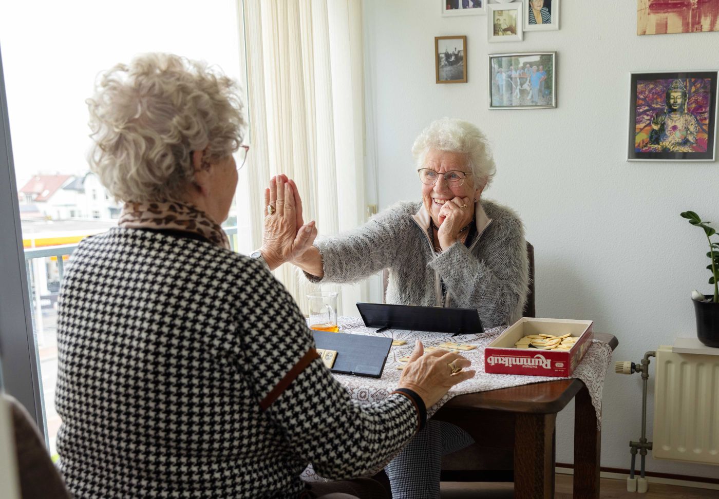 Cliënt en mantelzorger rummikub high five Oudere dames spelen een spelletje Rummikub aan tafel in de woonkamer en geven elkaar een high five