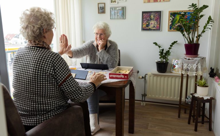 Cliënt en mantelzorger rummikub high five Oudere dames spelen een spelletje Rummikub aan tafel in de woonkamer en geven elkaar een high five