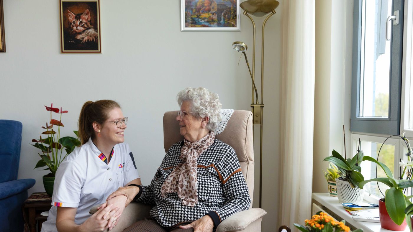 Cliënt in stoel in contact met zorgmedewerker woonkamer vacature thuiszorg Zorgmedewerker zit gehurkt naast oude mevrouw in stoel in de woonkamer in huis