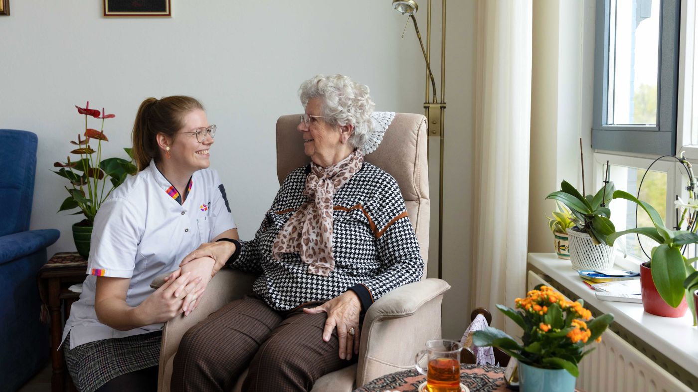 Cliënt in stoel in contact met zorgmedewerker woonkamer vacature thuiszorg Zorgmedewerker zit gehurkt naast oude mevrouw in stoel in de woonkamer in huis