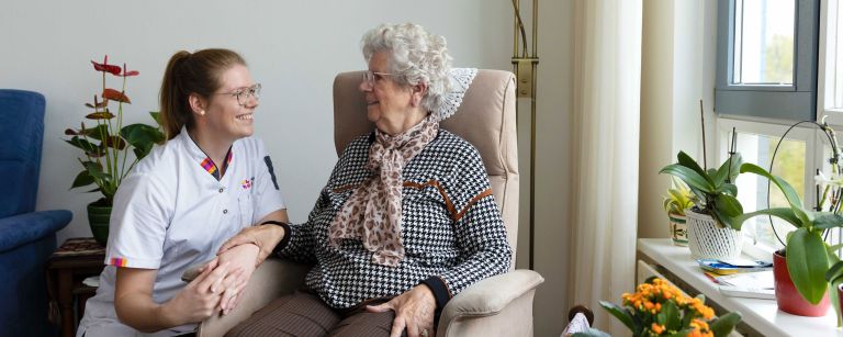 Cliënt in stoel in contact met zorgmedewerker woonkamer vacature thuiszorg Zorgmedewerker zit gehurkt naast oude mevrouw in stoel in de woonkamer in huis