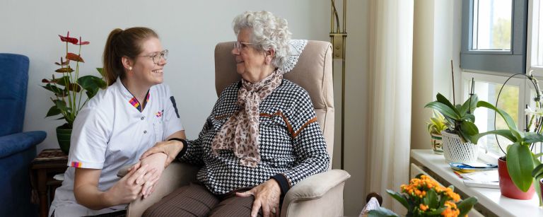 Cliënt in stoel in contact met zorgmedewerker woonkamer vacature thuiszorg Zorgmedewerker zit gehurkt naast oude mevrouw in stoel in de woonkamer in huis
