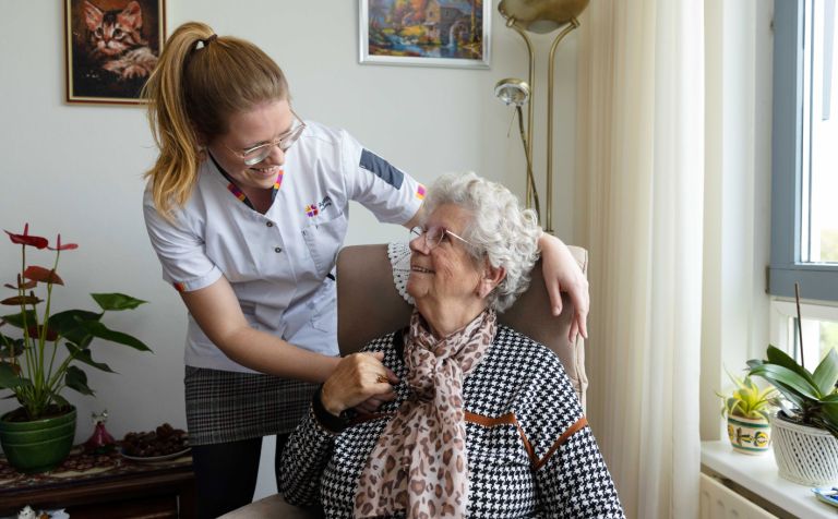 Cliënt in stoel met zorgmedewerker omhelzing woonkamer vacature thuiszorg Vrouwelijke zorgmedewerker omarmt een oude dame in een stoel in een woonkamer en kijkt de client aan