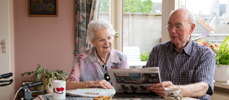Client echtpaar koffie krant tafel thuis Oud echtpaar drinkt koffie aan tafel in huis en leest de krant met rollator en alarmeringsknop