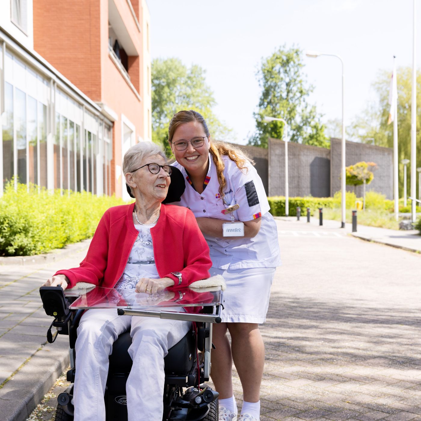 Client scootmobiel zorgmedewerker buiten vacature Riederborgh verpleeghuiszorg uniform Cliënt in scootmobiel rijdt buiten over straat met zorgmedewerker lachend erachter