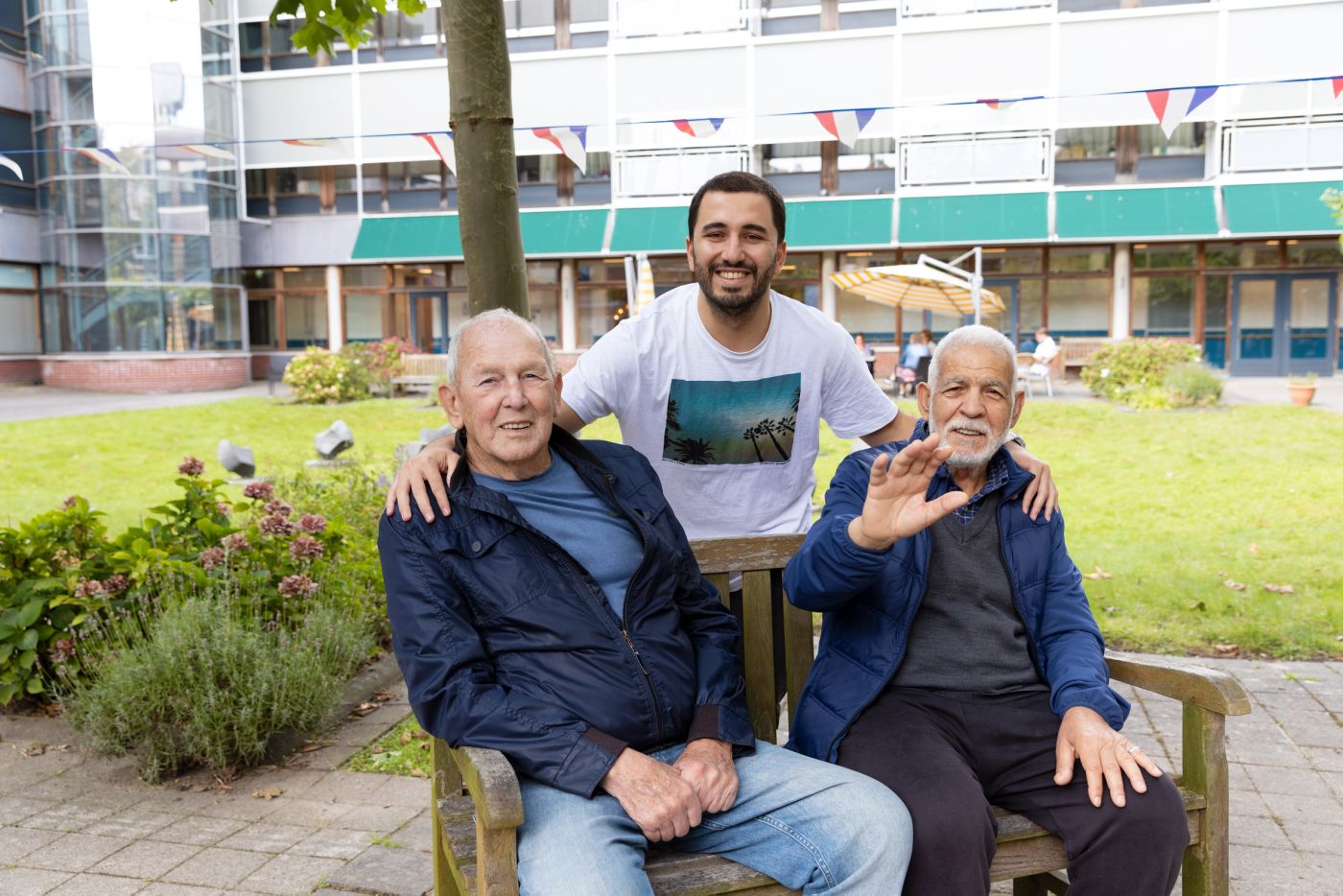Dagbesteding deelnemers begeleider tuin buiten - web Jonge meneer slaat zijn armen om twee oude mannen op een bankje in een tuin voor een verpleeghuis
