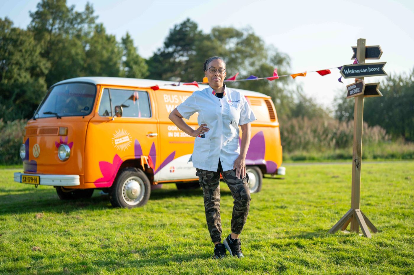 Medewerker Dania Blais christelijk Lelie zorggroep Siloam uniform bus vol waardering vacature verpleeghuiszorg zorg thuiszorg Vrouw met zorgjasje poseert voor oranje volkswagenbus in groene omgeving