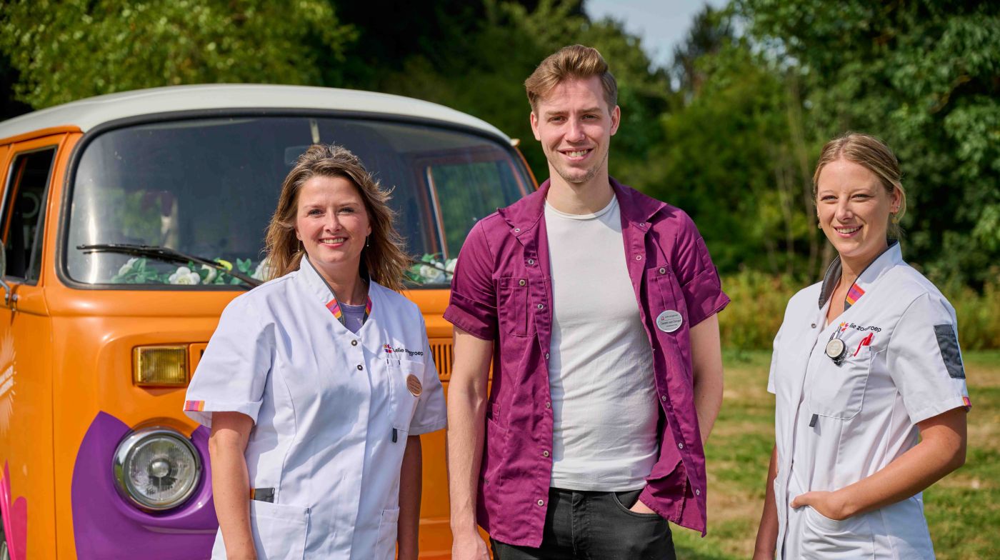 Drie zorgverleners voor bus van waardering van Lelie zorggroep Een man en twee vrouwen als zorgmedewerkers voor een oranje volkswagenbus van Lelie zorggroep