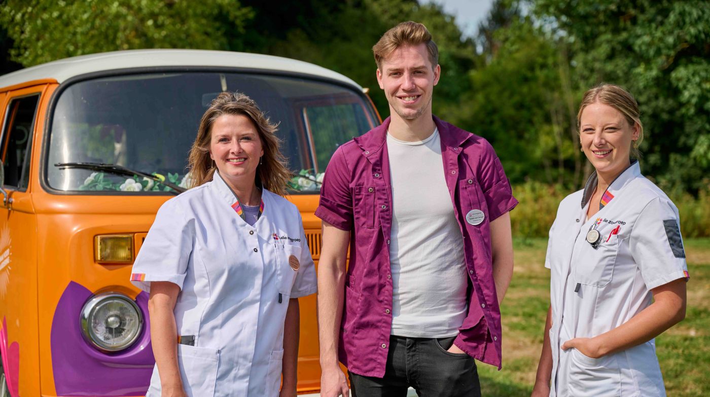 Drie zorgverleners voor bus vol waardering foto dichtbij vacature zorg Een man en twee vrouwen als zorgmedewerkers voor een oranje volkswagenbus van Lelie zorggroep