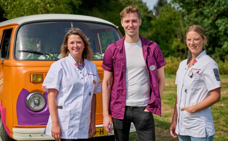 Drie zorgverleners voor bus vol waardering foto dichtbij vacature zorg Een man en twee vrouwen als zorgmedewerkers voor een oranje volkswagenbus van Lelie zorggroep