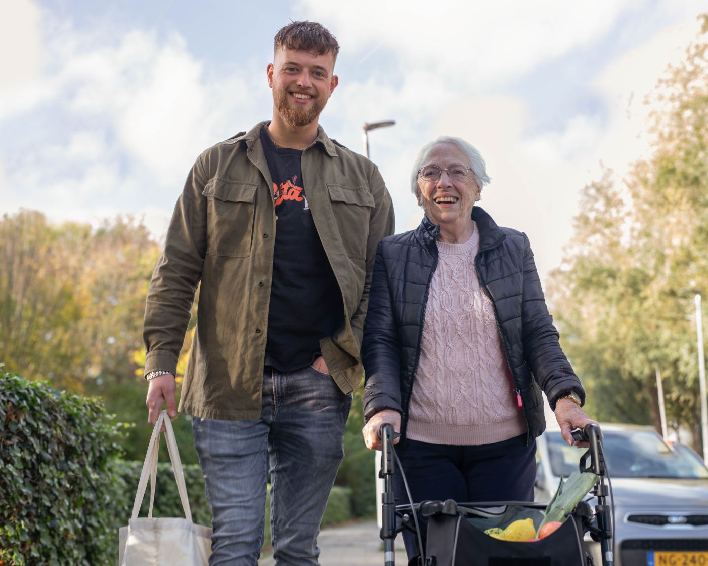 Bewoonster Siloam loopt met medewerker over straat met boodschappen Oudere dame achter rollator loopt met jonge man over straat met boodschappen