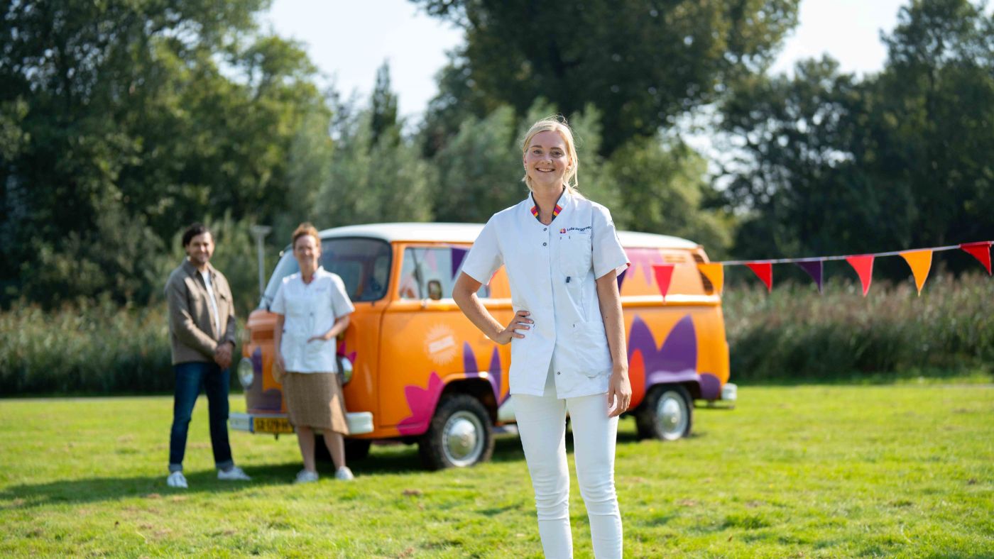 Vrouw van christelijke zorgorganisatie Lelie Zorggroep in Rotterdam vacature thuiszorg bus vol waardering verpleeghuiszorg uniform Jonge zorgmedewerker poseert glimlachend voor oranje vintage volkswagenbusje van Lelie zorggroep