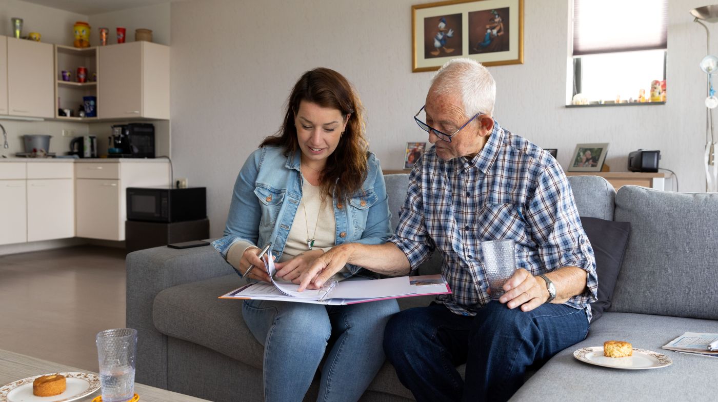 Hbh met client zorgdossier doornemen Vrouw hulp bij huishouden kijkt met mannelijke client naar documenten en zit op de bank
