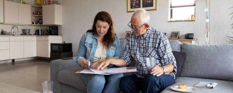 Hbh met client zorgdossier doornemen Vrouw hulp bij huishouden kijkt met mannelijke client naar documenten en zit op de bank