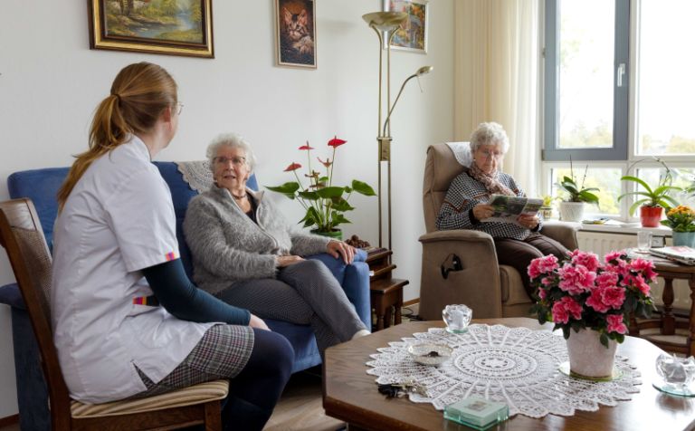 Huiskamer cliënt met mantelzorger en zorgmedewerker Zorgmedewerker zit in de huiskamer met twee oude vrouwen