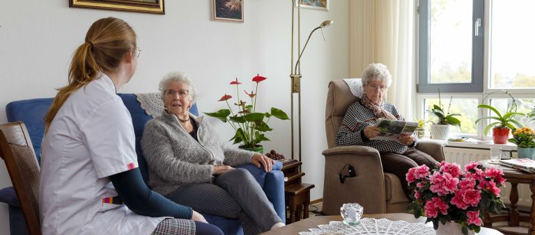 Huiskamer cliënt met mantelzorger en zorgmedewerker Vrouwelijke zorgmedewerker in gesprek met vrouwelijke client en mantelzorger op de bank in een woonkamer in huis