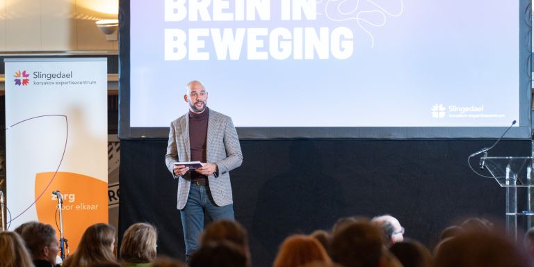 Presentatie van Korsakov expertisecentrum Slingedael Man in grijs net jasje staat op een podium naast een roll-up banner en presenteert met een microfoon in zijn hand