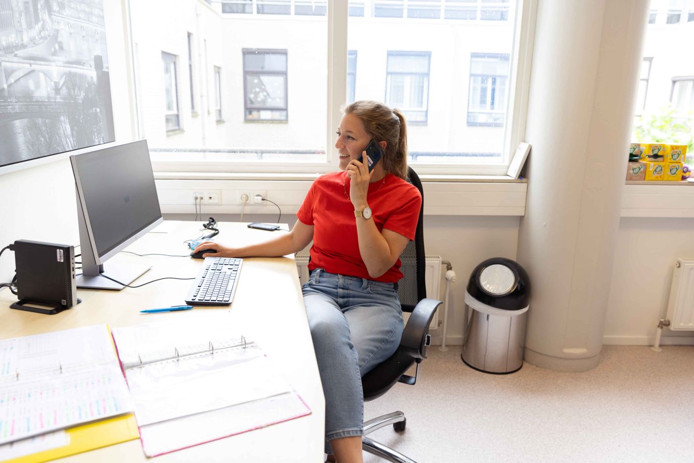 Medewerker van Lelie zorggroep achter computer bij Korsakov Expertisecentrum Slingedael in Rotterdam Vrouw zit achter bureau terwijl ze haar telefoon aan haar oor houdt en op het computerscherm kijkt met de muis in haar hand
