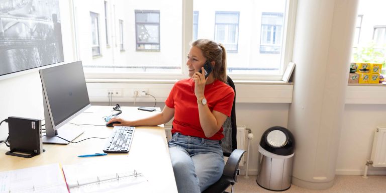 Medewerker van Lelie zorggroep achter computer bij Korsakov Expertisecentrum Slingedael in Rotterdam Vrouw zit achter bureau terwijl ze haar telefoon aan haar oor houdt en op het computerscherm kijkt met de muis in haar hand