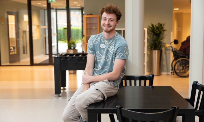 Sociaal pedagogisch medewerker van Korsakov Expertisecentrum Slingedael in Rotterdam Jonge man met rode krullen zit op een tafel in de ontmoetingsruimte van Slingedael en lacht in de camera