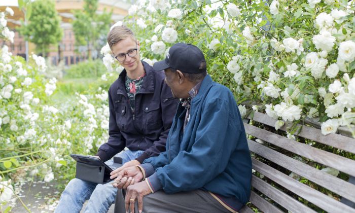 Cliënt en hulpverlener buiten op bank hulpverlening vacature gesprek zonderuniform behandeling psycholoog Meneer Pinas en jonge mannelijke hulpverlener Henri van Vliet zitten buiten op bank met witte bloemen voor ambulante hulpverlening
