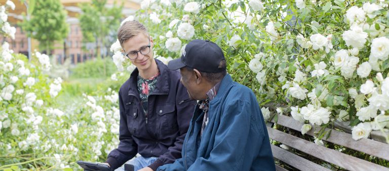Pinas en hulpverlener buiten op bank met witte bloemen-2 Jonge mannelijke hulpverlener en oudere meneer zitten buiten op een bank in een tuin in gesprek