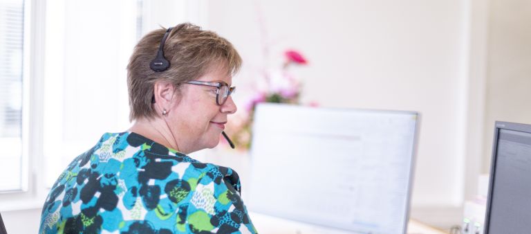 Receptioniste van christelijke zorgorganisatie Lelie zorggroep bellend achter computer bij de receptie vacature Vrouwelijke receptioniste van Lelie zorggroep zit achter computer en belt via headset