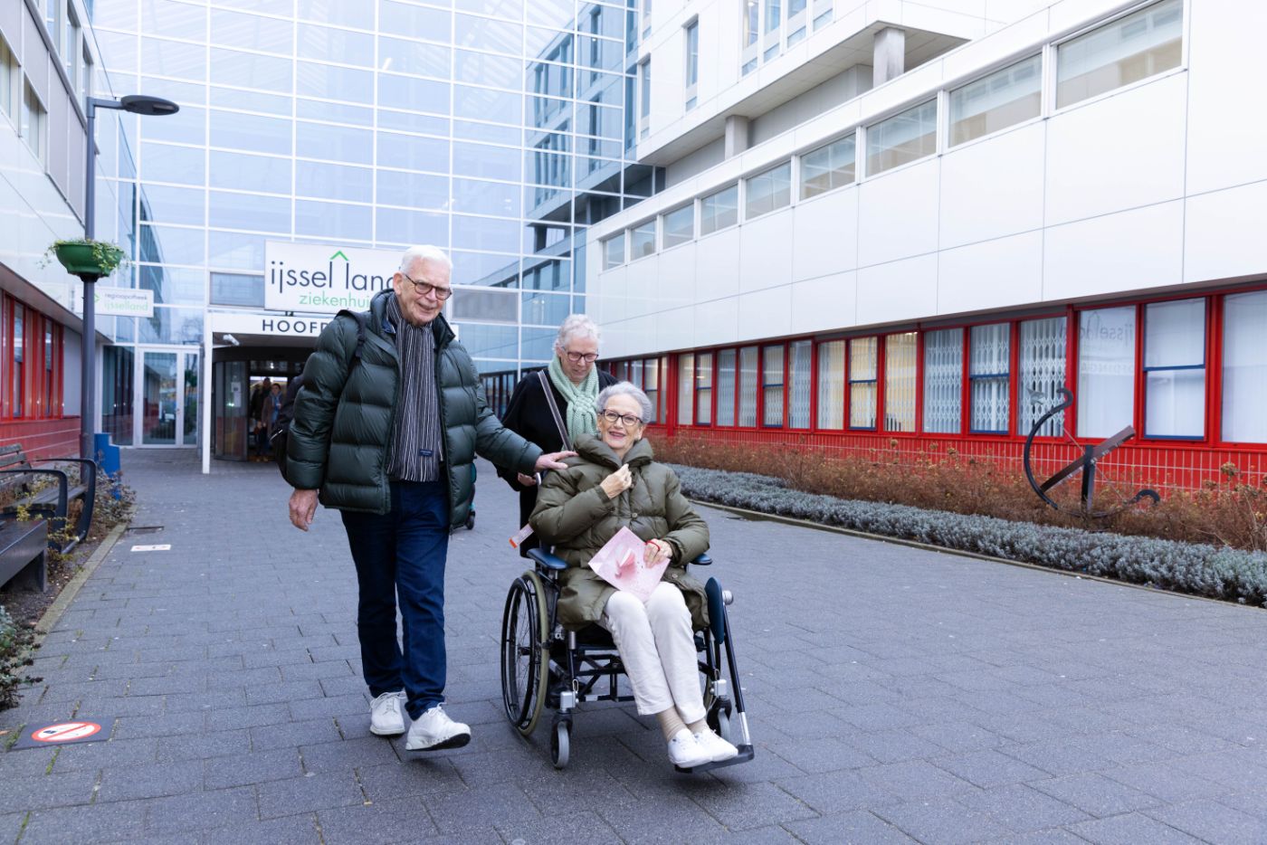 Revalidatie cliënt in rolstoel voor hoofdingang IJsselland Client zit in een rolstoel met familieleden om haar heen voor het IJsselland Ziekenhuis
