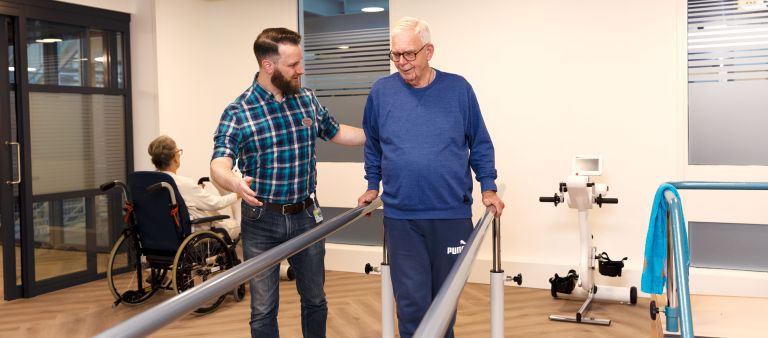 Revalidatie oefening lopen man met christelijke zorgverlener van Lelie zorggroep Een oudere man is bezig met een revalidatie oefening voor lopen tussen twee steunen zorgverlener staat ernaast