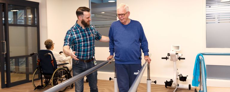 Revalidatie oefening lopen man met christelijke zorgverlener van Lelie zorggroep Een oudere man is bezig met een revalidatie oefening voor lopen tussen twee steunen zorgverlener staat ernaast