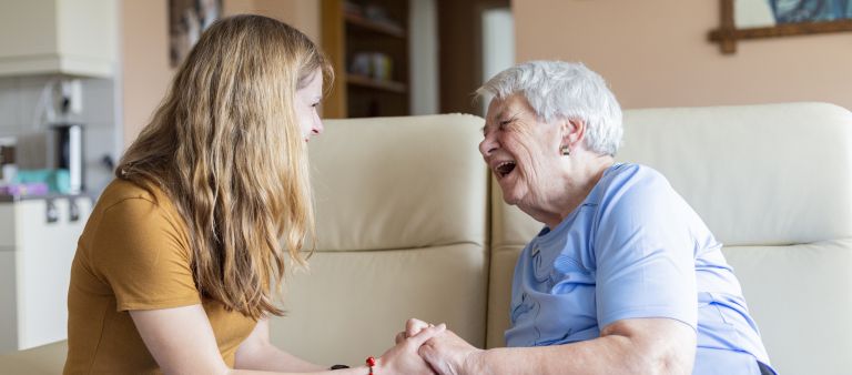 Logopedist en cliënt bij behandeling thuis van Lelie zorggroep in Rotterdam vacature welzijn logopedie dagbesteding Jonge vrouw en oude vrouw zitten op een bank in een huiskamer en houden lachend elkaars handen vast