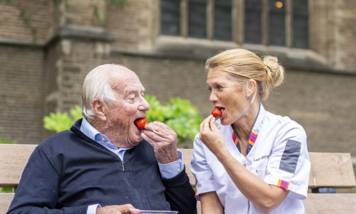 Diëtetiek en voedingsadvies bij Lelie zorggroep in Rotterdam, Ridderkerk en Hoogvliet voor behandeling thuis Oude man en vrouw in zorgjasje zitten tegenover elkaar op een stoel buiten en eten allebei een aardbei