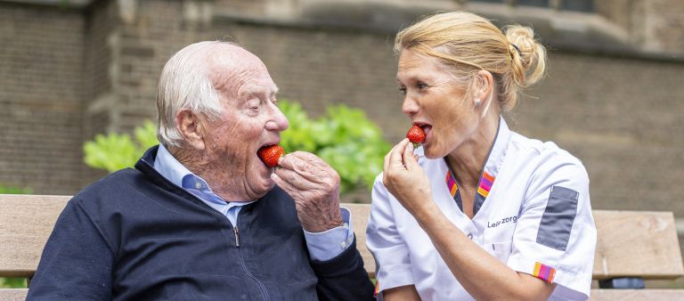 Diëtetiek en voedingsadvies bij Lelie zorggroep in Rotterdam, Ridderkerk en Hoogvliet voor behandeling thuis Oude man en vrouw in zorgjasje zitten tegenover elkaar op een stoel buiten en eten allebei een aardbei