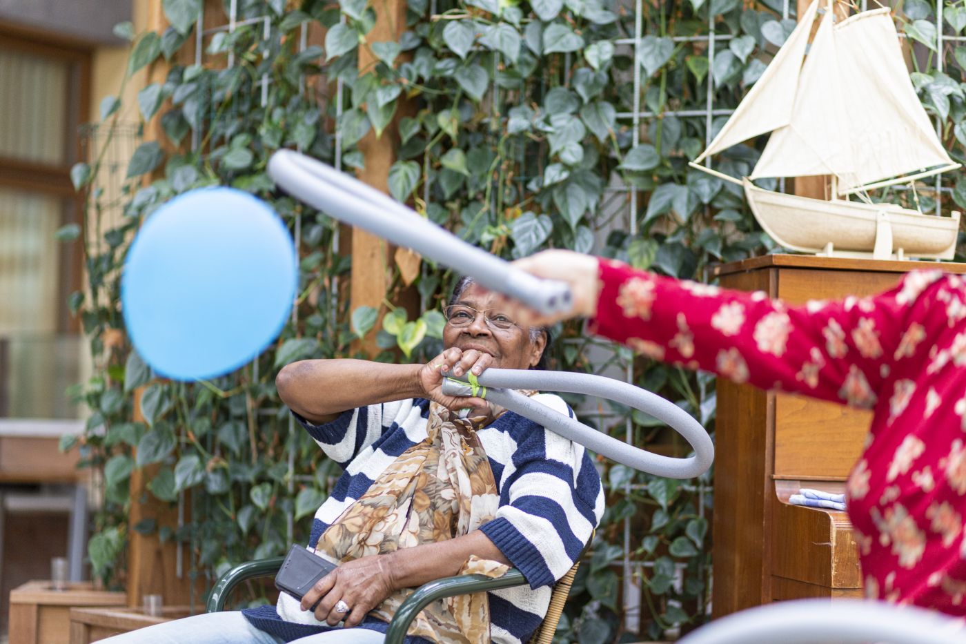 Vrouw in stoel voorgrond activiteit ballon slaan Oude mevrouw zit op een stoel en doet lachend een activiteit met ballonnen en rackets