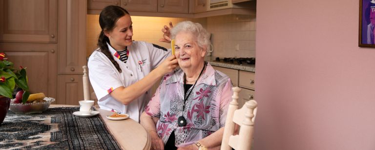 Zorgmedewerker client haar kammen thuiszorg keuken vacature thuiszorg uniform Vrouwelijke zorgmedewerker Lelie zorggroep kamt haar van oude vrouw bij tafel in de keuken