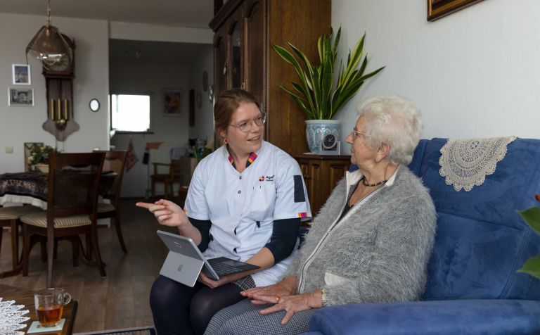 Zorgmedewerker en cliënt op bank in woonkamer met tablet Vrouwelijke zorgmedewerker en oude dame zitten op de bank in een woonkamer in huis en wijzen naar een tablet
