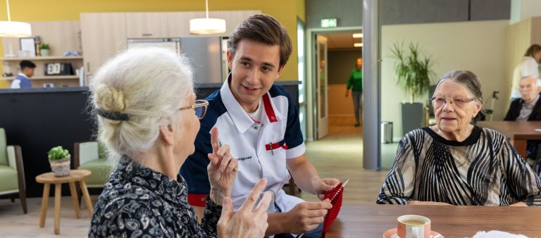 Zorgverlener en bewoner in woonkamer met breiwerk - web Mannelijke zorgverlener breit en is in gesprek samen met twee oude dames in het verpleeghuis op de huiskamer