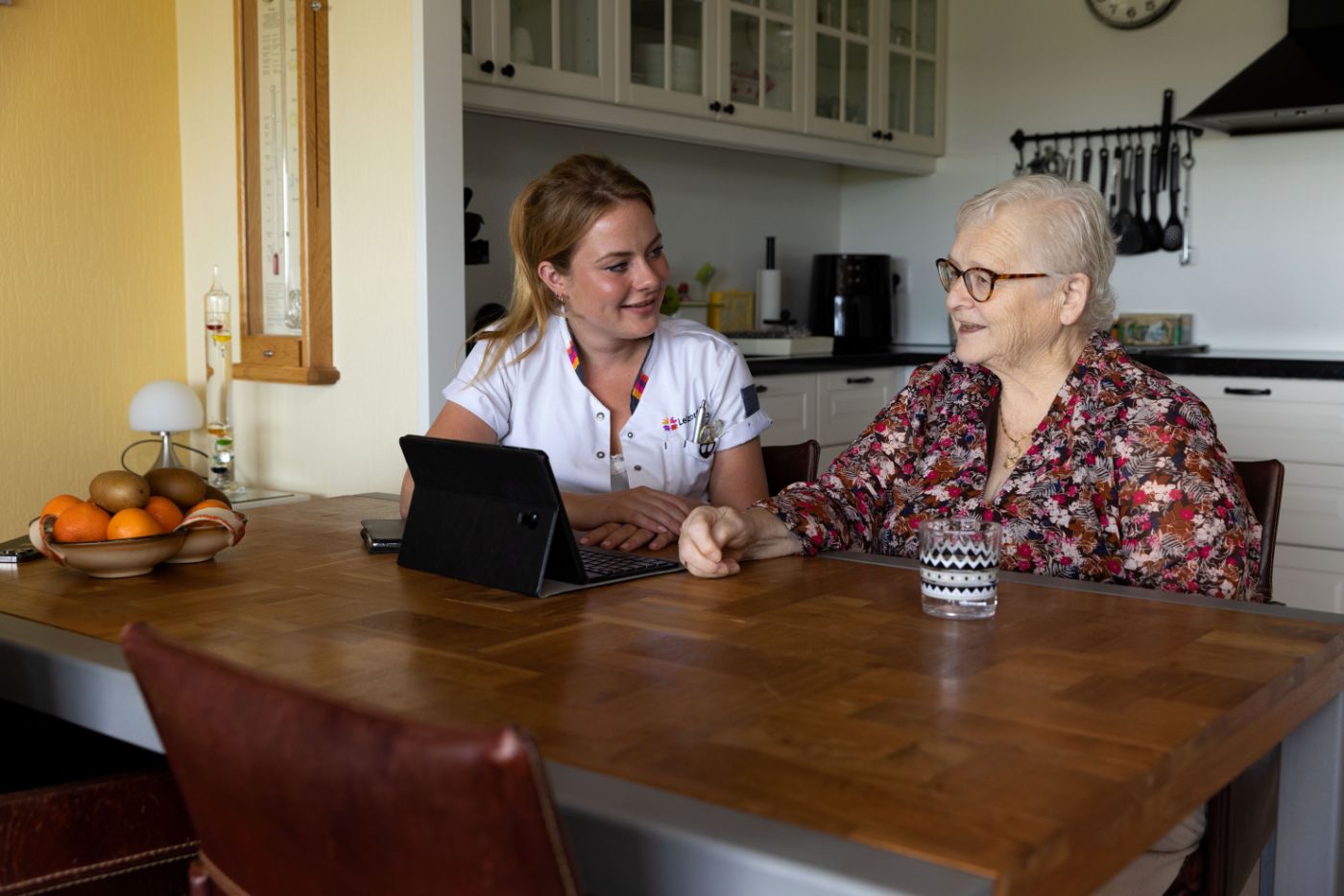 Zorgverlener en cliënt met tablet aan tafel Vrouw in zorgjasje en oude vrouw lachen naar elkaar en zitten samen aan tafel met een kopje koffie en een tablet