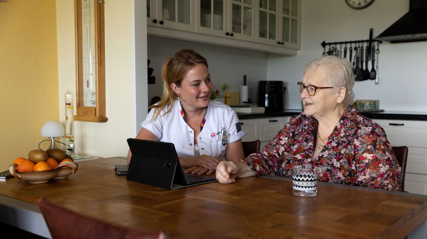 Christelijke jonge zorgverlener en cliënt met tablet aan tafel Jonge vrouwelijke zorgverlener zit te praten met de cliënt aan tafel achter een tablet