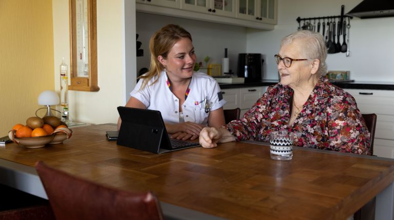 Christelijke jonge zorgverlener en cliënt met tablet aan tafel Jonge vrouwelijke zorgverlener zit te praten met de cliënt aan tafel achter een tablet