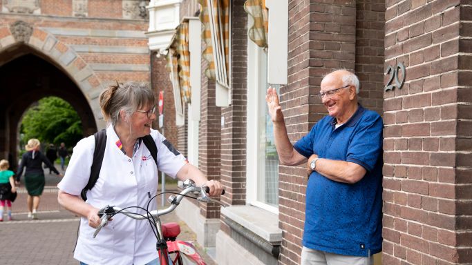 Zorgverlener fiets zwaaien client thuiszorg voordeur huis vacature thuiszorg Mannelijke client van middelbare leeftijd zwaait in de deuropening naar vrouwelijke zorgmedewerker buiten op de fiets