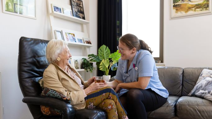 Bewoner en zorgverlener van Lelie zorggroep drinken thee Oude vrouw zit in een grote stoel met kopje thee en zorgverlener ernaast in de woonkamer