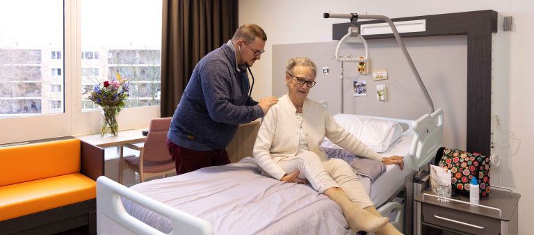 Specialist ouderengeneeskunde voor advies en diagnostiek bij behandeling thuis van Lelie zorggroep in Rotterdam, Ridderkerk en Krimpen aan den IJssel Een man staat in een ziekenhuiskamer bij een bed waar een vrouw op zit en hij onderzoekt haar met een stethoscoop