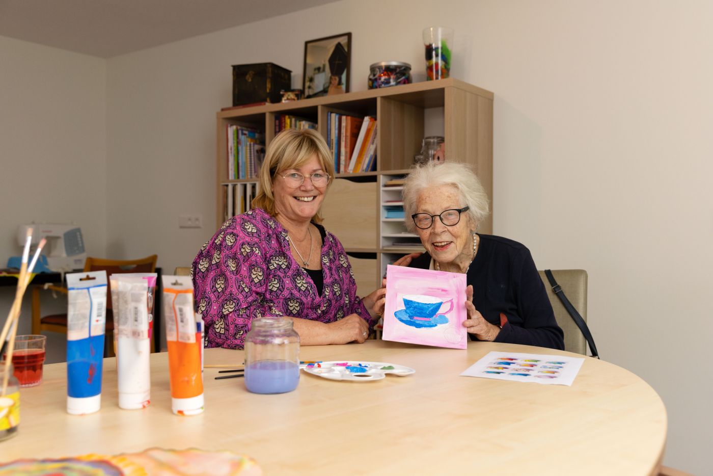 Deelnemer en begeleider van dagbesteding Carmenhof schilderen bij christelijke zorgorganisatie Lelie zorggroep in Rotterdam vacature welzijn vrijwilliger Jonge vrouw en oudere vrouw zitten samen aan creatieve tafel en oude vrouw houdt haar schilderwerk omhoog