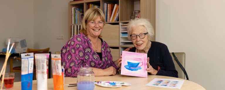 Deelnemer en begeleider van dagbesteding Carmenhof schilderen bij christelijke zorgorganisatie Lelie zorggroep in Rotterdam Jonge vrouw en oudere vrouw zitten samen aan creatieve tafel en oude vrouw houdt haar schilderwerk omhoog