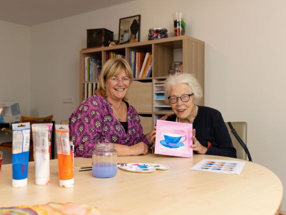 Deelnemer en begeleider van dagbesteding Carmenhof schilderen bij christelijke zorgorganisatie Lelie zorggroep in Rotterdam vacature welzijn vrijwilliger Jonge vrouw en oudere vrouw zitten samen aan creatieve tafel en oude vrouw houdt haar schilderwerk omhoog