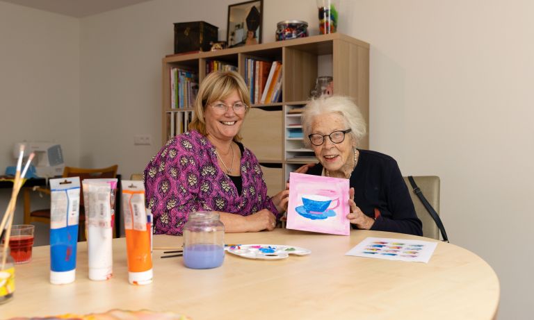 Deelnemer en begeleider van dagbesteding Carmenhof schilderen bij christelijke zorgorganisatie Lelie zorggroep in Rotterdam vacature welzijn vrijwilliger Jonge vrouw en oudere vrouw zitten samen aan creatieve tafel en oude vrouw houdt haar schilderwerk omhoog