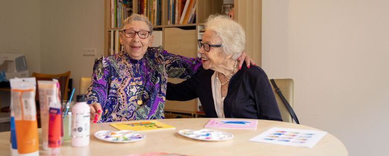 Twee deelnemers van de dagbesteding van christelijke zorgorganisatie Lelie zorggroep in Rotterdam bij het schilderen Twee oude vrouwen zitten aan een tafel de vol staat met creatieve schilderspullen en ze lachen met elkaar