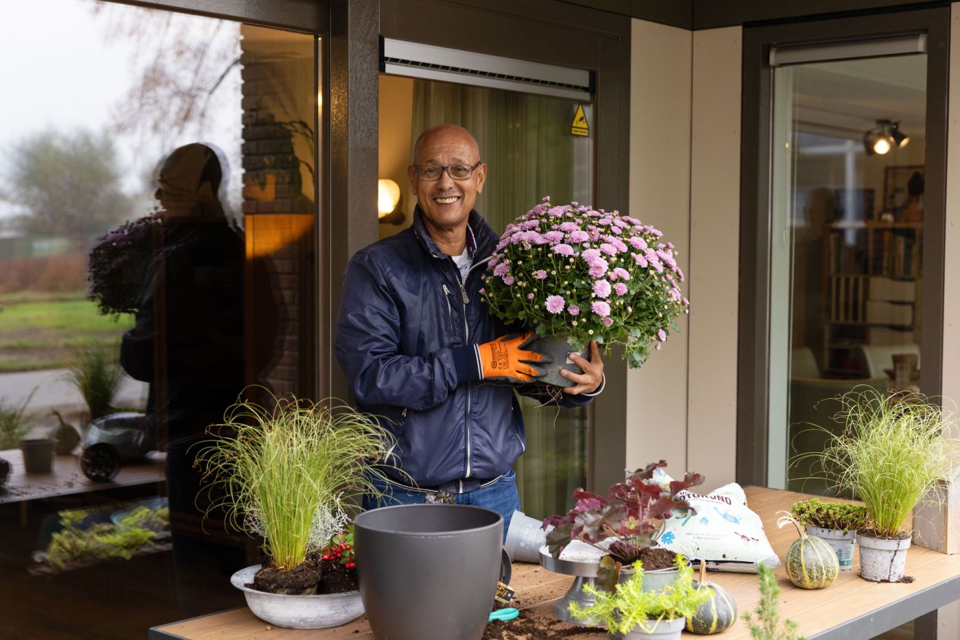 Deelnemer dagbesteding christelijke zorgorganisatie Lelie zorggroep tuinieren vacature vrijwilliger facilitair welzijn Man staat buiten bij tuintafel met allerlei spullen voor tuinieren en hij houdt lachend een mooie plant omhoog