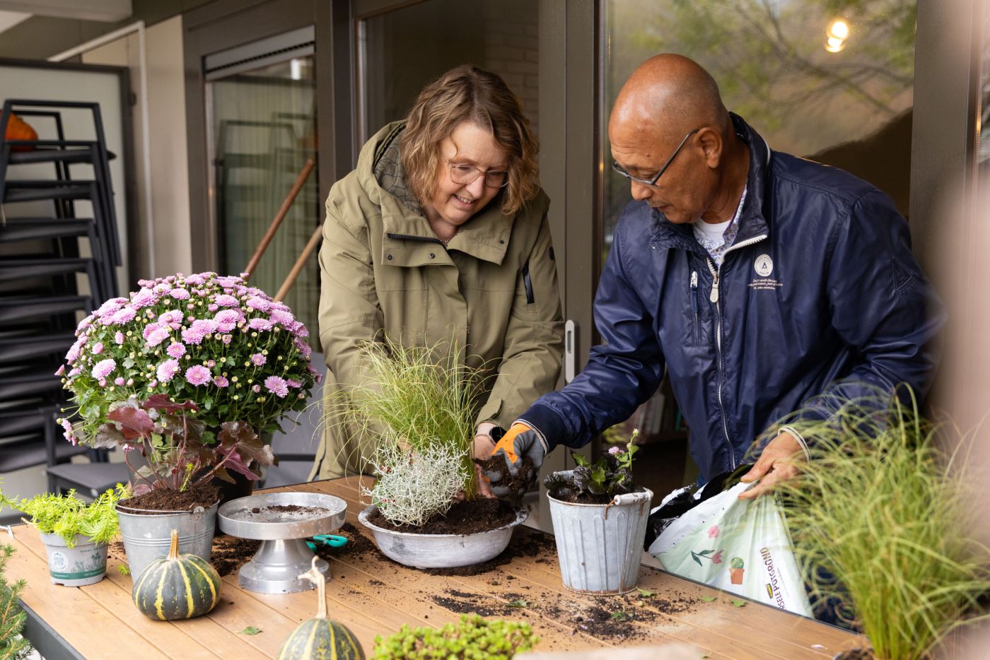 Begeleider en deelnemer van de dagbesteding van christelijke zorgorganisatie Lelie zorggroep in Rotterdam vacature dagbesteding tuinieren deelnemer en begeleider welzijn Een vrouw en een man staan buiten bij een tuintafel en stoppen samen potgrond in een pot